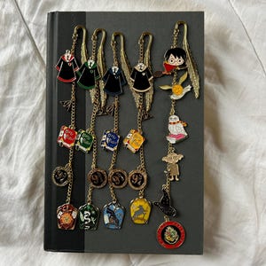 May include: A collection of gold-toned metal Harry Potter-themed bookmarks. Each bookmark features a charm with various designs, including Hogwarts house robes, Harry Potter on a broom, and other iconic symbols from the series. The bookmarks are displayed on a closed book.