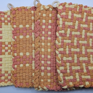 May include: Four woven potholders in shades of peach, orange, and cream. Each potholder has a square shape with a woven pattern and a wooden ring for hanging. The edges are finished with a twisted rope trim.