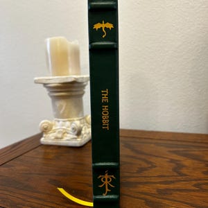 Puede incluir: Un libro de tapa dura verde con un drag&oacute;n dorado y el t&iacute;tulo "THE HOBBIT" en el lomo. Un marcap&aacute;ginas dorado con un s&iacute;mbolo est&aacute; unido al libro.