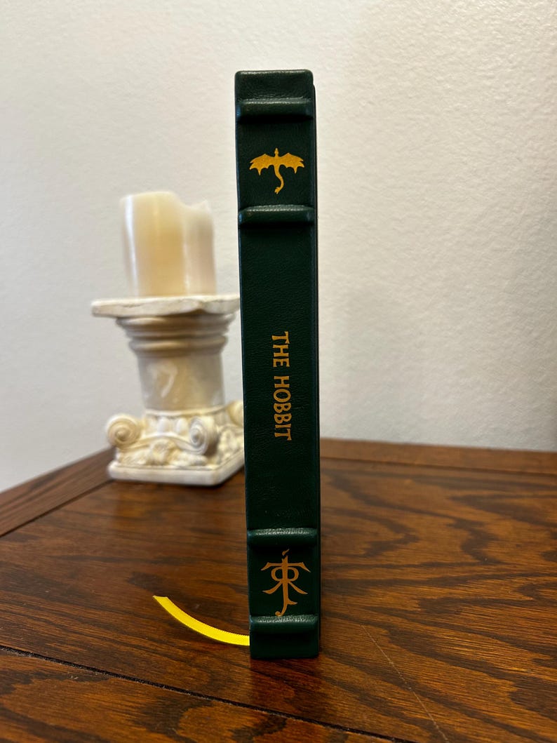 Puede incluir: Un libro de tapa dura verde con un drag&oacute;n dorado y el t&iacute;tulo "THE HOBBIT" en el lomo. Un marcap&aacute;ginas dorado con un s&iacute;mbolo est&aacute; unido al libro.