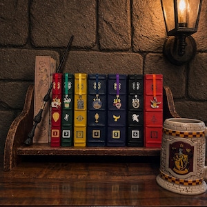 May include: A collection of seven colorful, numbered book-shaped boxes with decorative charms and a wand on a wooden shelf. A ceramic mug with a crest sits to the right. The background features a brick wall and a wall-mounted lantern.