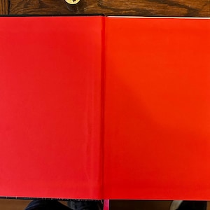 以下が含まれることがあります： 赤いページが開かれた本。左ページは右ページよりも濃い赤色です。本は黒い表紙とピンクのリボンのしおりが付いています。本は木製の表面に置かれています。