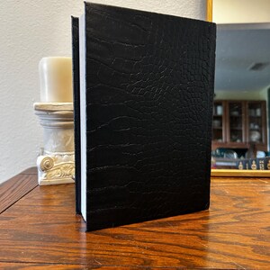 以下が含まれることがあります： 木製の表面に、クロコダイルのような模様の黒い本が立っています。本のページは白く、背表紙が見えます。白いろうそくと装飾的なスタンドが背景にあります。