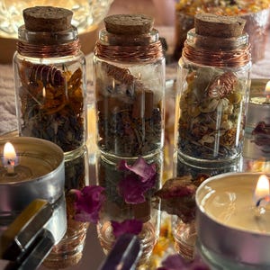 May include: Three small glass jars with cork stoppers, filled with dried herbs and spices, are arranged on a reflective surface. The jars are wrapped with copper wire. Lit candles and crystals surround the jars, creating a mystical aesthetic.