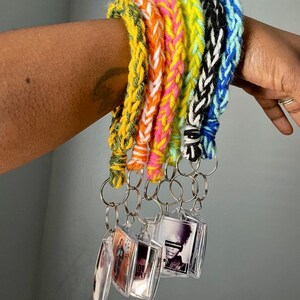 May include: A collection of five braided yarn wristlet keychains in yellow, orange, pink, lime green, and black and blue. Each keychain features a clear acrylic rectangular frame containing a small black and white photograph.