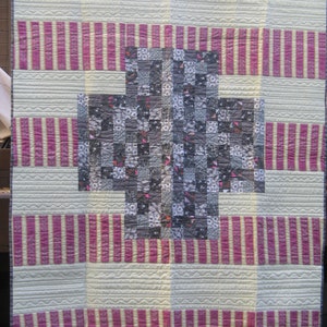 May include: A quilt with a large, central, patchwork design in shades of gray, black, and white. The patchwork is surrounded by a border of alternating pink and white stripes.