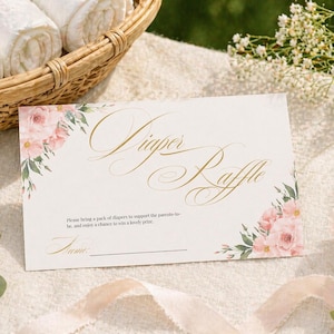 Könnte beinhalten: Eine weiße Karte mit goldener Schrift "Diaper Raffle", verziert mit rosa floralen Akzenten. Die Karte liegt auf einer weißen Oberfläche, im Hintergrund ein Korb mit gerollten weißen Handtüchern.