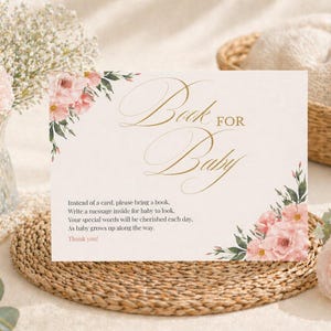 Könnte beinhalten: Eine Babyparty-Einladungskarte mit den Worten "Book for Baby" in eleganter goldener Schrift. Die Karte ist mit Aquarell-Blumenakzenten verziert und enthält eine Nachricht, in der um ein Buch anstelle einer Karte gebeten wird.