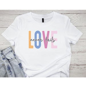 May include: White t-shirt featuring the phrase "LOVE never fails" in colourful block letters. The word "LOVE" is in orange, blue, and pink, with "never fails" in black script. The shirt is shown with blue jeans.