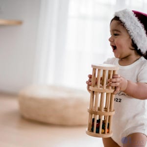 May include: A baby wearing a red and white Santa hat holds a wooden toy with colorful beads inside. The toy has a cylindrical shape with multiple rings. The baby is wearing a white onesie with the number 22 printed on it.