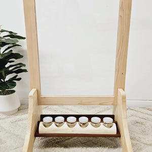 Könnte beinhalten: Ein Holzstaffelei mit einer durchsichtigen Acrylglasscheibe und einem Regal für kleine Behälter. Die Staffelei steht auf einem hellbeigen Teppich.