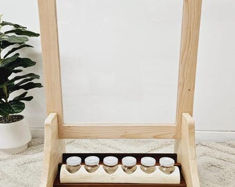 May include: A wooden easel with a clear acrylic panel and a shelf for small containers. The easel is standing on a light beige rug.