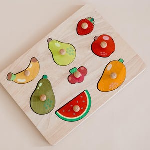 May include: Wooden puzzle featuring various fruits, including a banana, pear, avocado, watermelon, mango, and strawberries. The puzzle pieces are brightly colored and have a knob on the back for easy handling.