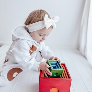 Puede incluir: Un niño juega con un juguete de apilar de madera de colores. El juguete está hecho de bloques de diferentes colores que encajan entre sí. Los bloques son rojo, naranja, amarillo, verde, azul y morado. El niño lleva una diadema blanca con un lazo.