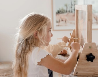May include: A young girl uses a wooden easel with a clear glass surface to paint. The easel has a knob on the side and is made of natural wood.