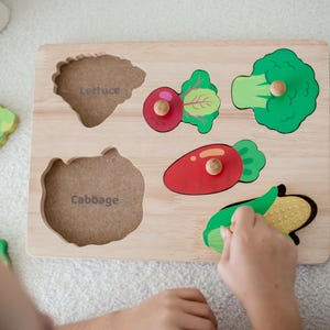 May include: A wooden puzzle featuring colourful vegetable shapes, including a green lettuce, a red beetroot, a green broccoli, a red carrot, a green corn, and a green cabbage. The puzzle pieces are labelled with the names of the vegetables.