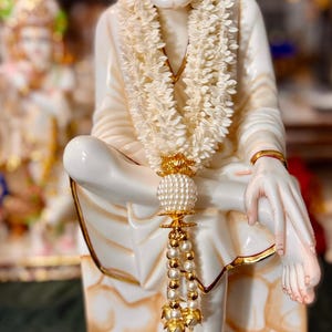 May include: A white ceramic statue of a seated spiritual figure, adorned with a garland and gold accents. The statue has a serene expression and is seated on a pedestal. Other statues are visible in the background.