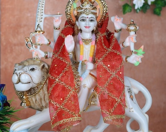 May include: A white marble statue of a deity riding a lion. The deity wears a red shawl with gold accents and a golden crown. The statue has multiple arms and is adorned with jewelry. The lion is also white with gold details.