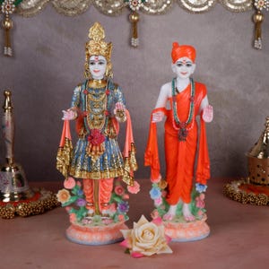 May include: Two statues of religious figures. One figure is dressed in blue and gold clothing, the other in orange. Both figures stand on pedestals decorated with flowers. A rose is in the foreground.