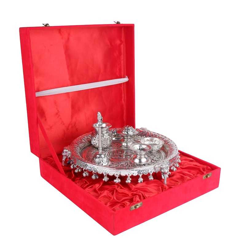 Puede incluir: Un juego de thali de puja plateado con detalles ornamentados, que incluye peque&ntilde;os cuencos y un recipiente decorativo, presentado en una caja forrada de terciopelo rojo. El thali es redondo y descansa sobre peque&ntilde;os pies.