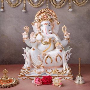 May include: A white and gold statue of Ganesha, seated on a lotus flower, with intricate detailing. The statue has a gold halo and is adorned with gold accents. A small bell and flowers are in the foreground, with decorative garlands in the background.