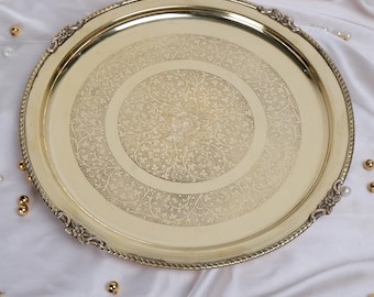 May include: A round, gold-colored metal tray with an ornate floral design etched into the center. The tray has a decorative rope-like edge and decorative corner accents. The tray is approximately 14 inches in diameter.