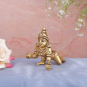 May include: A golden-colored statue of a crawling figure, possibly a deity, with intricate details in its clothing and headdress. The statue is on a blue surface, with a pink and white background and a white rose.
