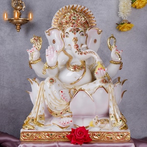 Puede incluir: Estatua blanca y dorada de Ganesha, deidad hindú, sentado sobre una flor de loto. La estatua presenta intrincados detalles dorados, detalles rojos y verdes y una corona decorativa. Una rosa roja se encuentra frente a la estatua.