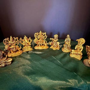 Op de afbeelding: Een verzameling goudkleurige religieuze beelden, waaronder afbeeldingen van goden. De beelden zijn gerangschikt op een groene doek, met een donkere achtergrond. De beelden variëren in grootte en pose en tonen ingewikkelde details.