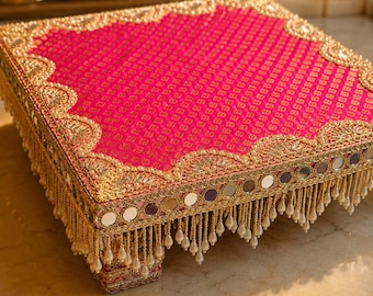 Puede incluir: Una plataforma cuadrada elevada cubierta con tela de color rosa intenso con un patrón texturizado. Los bordes están adornados con intrincados bordados dorados, espejos circulares y un fleco colgante de pequeñas perlas blancas.