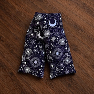 May include: A rectangular fabric heating pad featuring a dark navy blue background with a repeating pattern of white crescent moons and various starburst designs. The item is folded in half, displaying the celestial print across its entire surface.