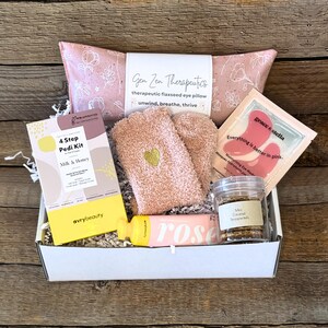 May include: A gift box filled with self-care items. Includes a pink eye pillow, fuzzy socks with a gold heart, a pedi kit, a rose-scented product, and a jar of caramel popcorn. The box is on a wooden surface.