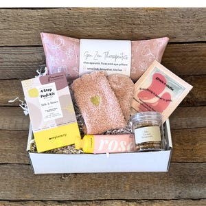 May include: A gift box filled with self-care items. Includes a pink eye pillow, fuzzy socks with a gold heart, a pedi kit, a rose-scented product, and a jar of caramel popcorn. The box is on a wooden surface.