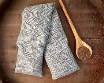 May include: A gray and white patterned heating pad rests on a wooden surface, next to a wooden spoon filled with brown seeds. The heating pad has a textured, woven design. The spoon is made of light-colored wood.