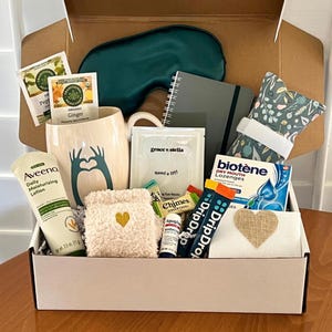May include: A gift box containing an Aveeno Daily Moisturizing Lotion 2.5 oz tube, a cream mug with a teal heart-hand graphic, a dark green sleep mask, a gray notebook, a floral eye pillow, Biotene lozenges, DripDrop packets, and Chimes ginger chews.