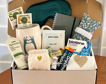 May include: A gift box containing an Aveeno Daily Moisturizing Lotion 2.5 oz tube, a cream mug with a teal heart-hand graphic, a dark green sleep mask, a gray notebook, a floral eye pillow, Biotene lozenges, DripDrop packets, and Chimes ginger chews.
