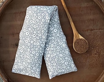 May include: A gray floral-patterned heat pack, folded in half, rests on a wooden tray. The heat pack is covered in small white flowers with black centers. A wooden spoon filled with seeds sits nearby.