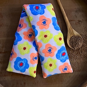 May include: A floral-patterned heat pack in a light gray fabric. The flowers are in shades of blue, orange, and yellow with red and blue centers. A wooden spoon filled with seeds rests beside the pack on a wooden tray.