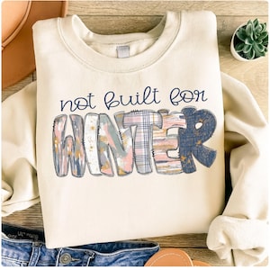 May include: Cream-colored sweatshirt with the phrase "not built for WINTER" in navy blue and a colorful, patterned design. The word WINTER is in a mix of plaid, pink, and blue patterns. A casual, cozy winter-themed apparel item.