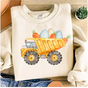 Könnte beinhalten: Cremefarbenes Sweatshirt mit einem gelben Muldenkipper, der mit Ostereiern und Karotten gefüllt ist. Der LKW hat schwarze Räder und ein detailliertes Design. Die Grafik ist mittig auf der Vorderseite des langärmeligen Oberteils angebracht.