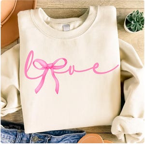 Peut inclure: Sweat-shirt crème avec le mot "Love" en script rose, orné d'un motif de nœud. Le sweat-shirt est plié, avec les manches visibles. L'image comprend un jean bleu et une sandale en cuir marron.