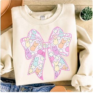May include: Cream-colored sweatshirt featuring a large, colorful bow design. The bow is decorated with pastel-colored bunny figures and white flowers on a pink background. The sweatshirt is a casual, comfortable style.