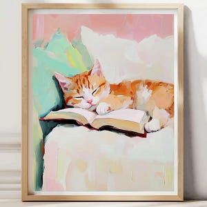May include: A framed art print depicts a sleeping orange tabby cat resting on an open book. The cat is lying on a white surface with a light green and pink background. The artwork is in a painterly style.