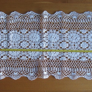 May include: White crocheted table runner with a scalloped edge and a delicate floral pattern. The runner measures approximately 122 cm long.