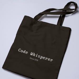 May include: A black tote bag with the text "Code Whisperer Since 2019" printed in white. The bag has two long handles for carrying over the shoulder or by hand. The bag is made of a durable material.