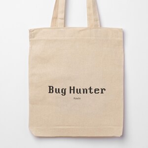 May include: A beige tote bag featuring the text "Bug Hunter" in a pixelated font. The name "Kevin" is printed below. The bag has two handles and is made of a light-coloured fabric.