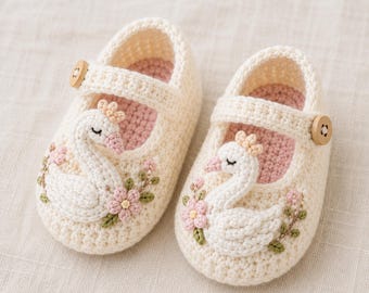 Peut inclure: Une paire de chaussons pour bébé en crochet couleur crème avec une application de cygne sur le devant. Chaque chausson possède une bride avec un petit bouton en bois, une doublure intérieure rose pâle et des broderies de fleurs roses et feuilles vertes.