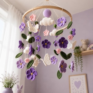 Puede incluir: Un móvil colgante para habitación infantil con elementos de ganchillo suspendidos de un marco circular de madera. Incluye flores moradas y rosas, mariposas blancas, nubes blancas y hojas verdes, todo elaborado con hilo y cuerdas blancas.