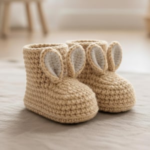 Puede incluir: Un par de botines de bebé de ganchillo, de color marrón claro. Cada botín presenta un diseño de orejas de conejo con un interior beige claro. Los botines están hechos de un hilo suave y texturizado, diseñados para la comodidad y el calor.