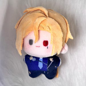 May include: A plush doll with fair skin, blonde hair, and a blue suit. The doll has a small red mark on its face and black eyes. The doll is made of soft fabric and is designed to resemble a character.